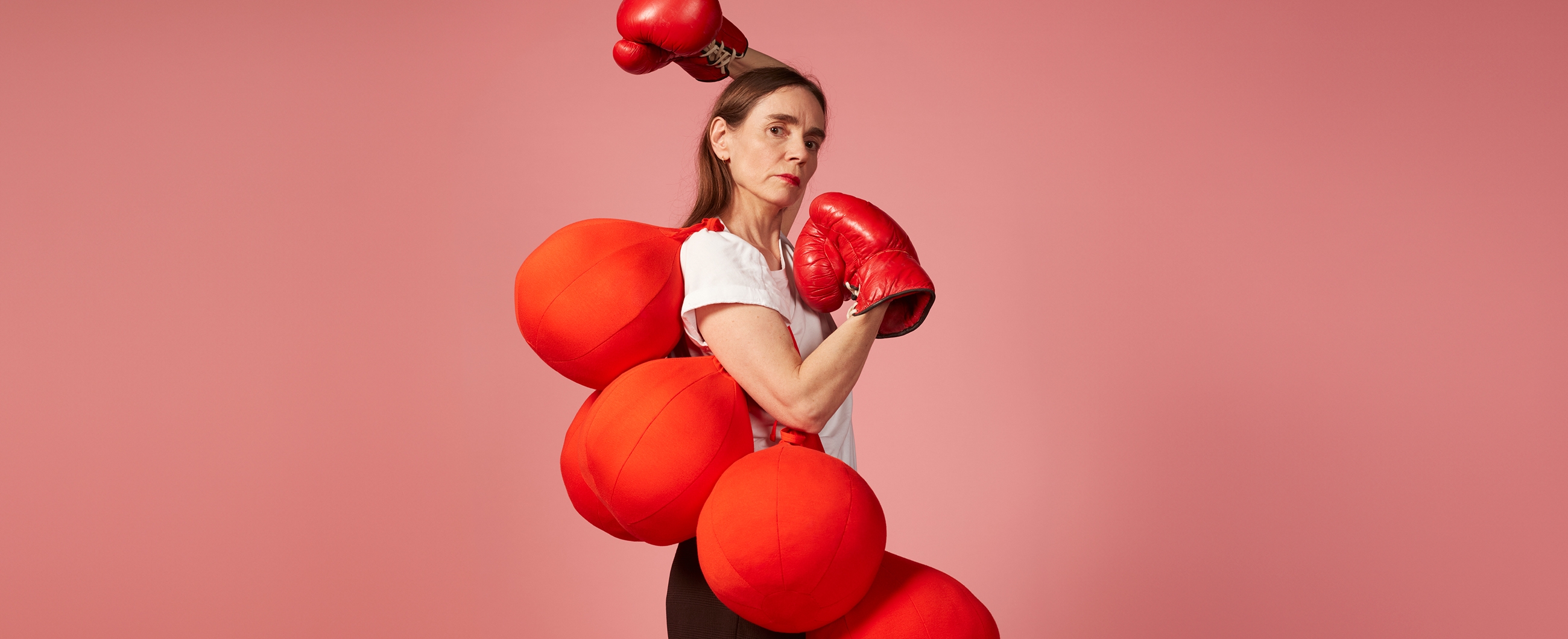 Eine Schauspielerin mit sehr grossen Boxhandschuhen trägt grosse Bälle über der Schulter. Sie blickt streng in die Kamera.